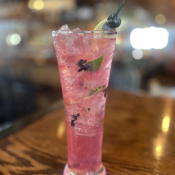 Pink cocktail with ice, lime, blueberries, and mint, on a wooden table at La Taguara Bar & Grill.