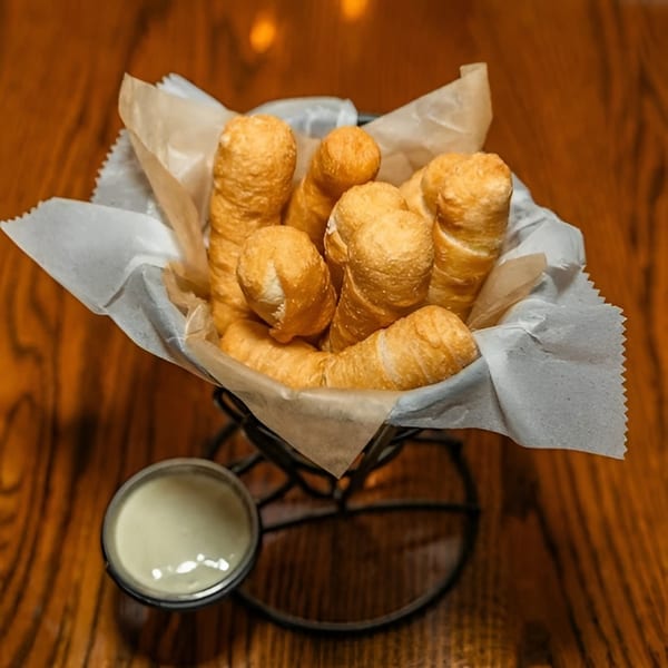 A basket of golden-brown Venezuelan tequeños, with a side of creamy white dipping sauce on a wooden table.