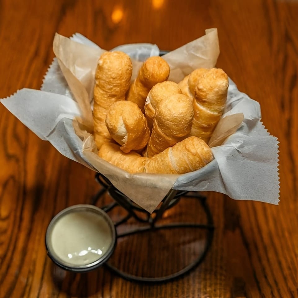 A basket of golden-brown Venezuelan tequeños, with a side of creamy white dipping sauce on a wooden table.