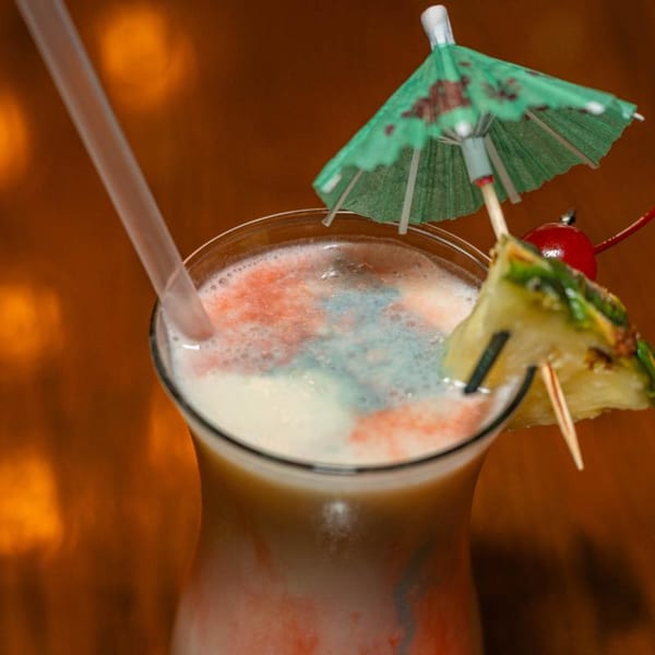 Colorful layered cocktail with a straw, pineapple, cherry, and a small green umbrella.