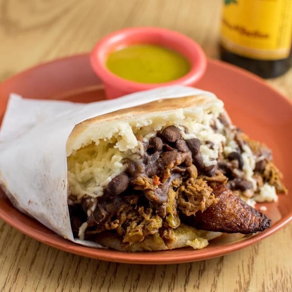 Venezuelan arepa stuffed with shredded beef, black beans, rice, cheese, and plantains, with green sauce.