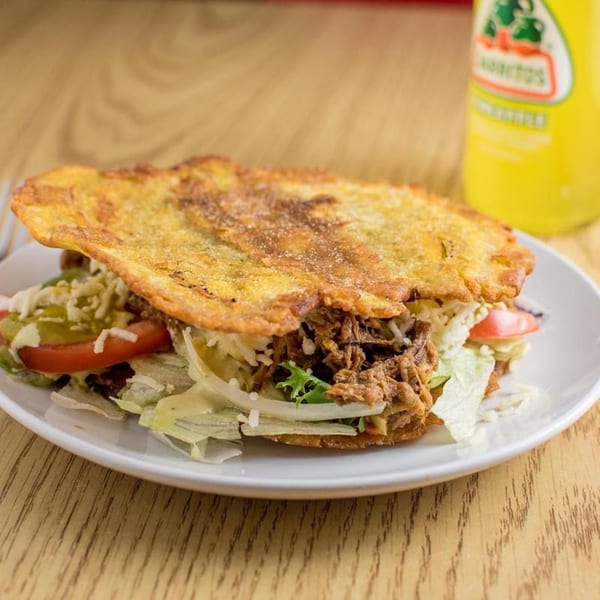 A Venezuelan patacon with shredded beef, lettuce, tomato, and cheese, next to a yellow soda.