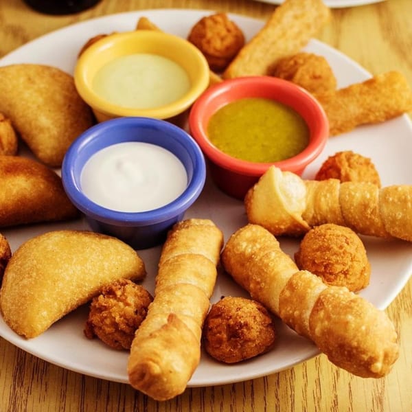 A plate of Venezuelan appetizers including empanadas, tequeños, and mandocas with three dipping sauces.