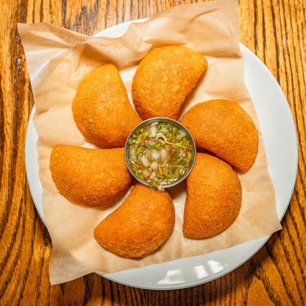 Six golden-fried Venezuelan empanadas arranged around a small bowl of green salsa on a white plate.